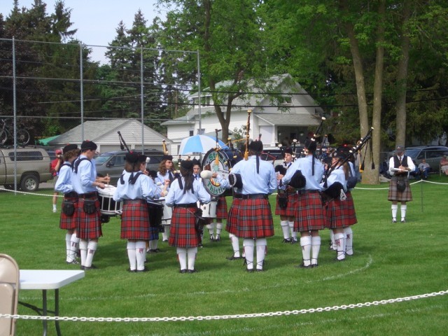 Pipe band in red tartan kilts playing in a circle on a lawn in Alma